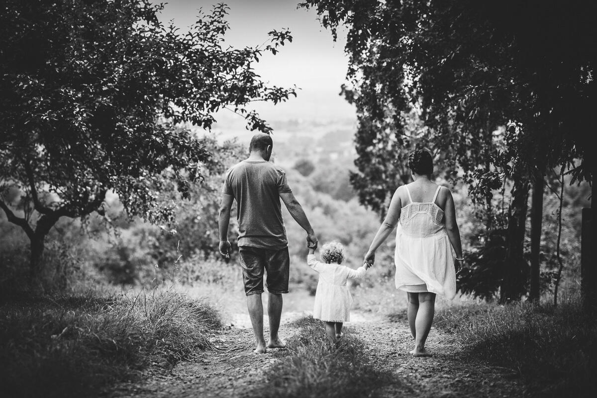 Wspólna droga – czarno-białe kadry pełne emocji Czarno-biała fotografia rodzinna w lesie – rodzice prowadzący dwuletnią dziewczynkę leśną drogą, ujęcie od tyłu, naturalny spacer na sesji w Krakowie.